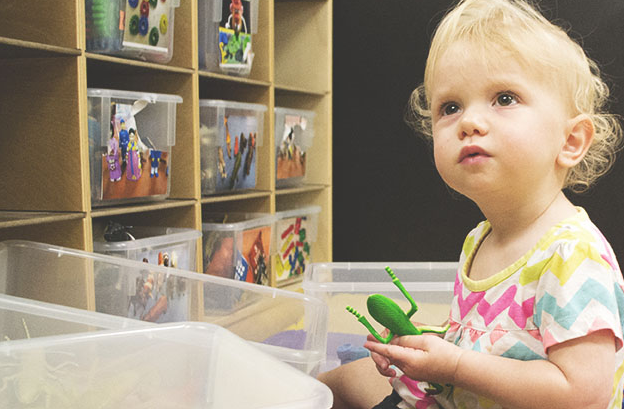 A toddler playing with a plastic grasshopper by the toy shelf looks up.